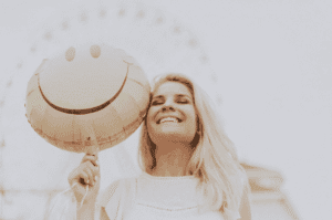 Jeune femme blonde, aux cheveux longs, souriante tenant un ballon smiley à la fête foraine.