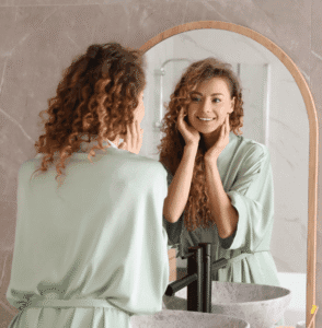 Femme aux cheveux bouclés se regardant dans un miroir, concentrée, heureuse.