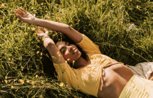Femme souriante allongée dans l’herbe, se reposant. Elle porte un chemisier et un bas jaunes.